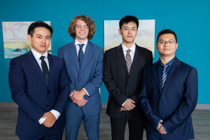 Group of four students in suits standing in front of blue wall