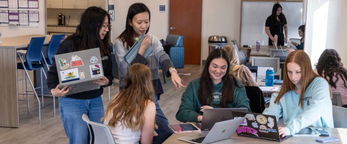 Students at a table and other students standing pointing at a laptop